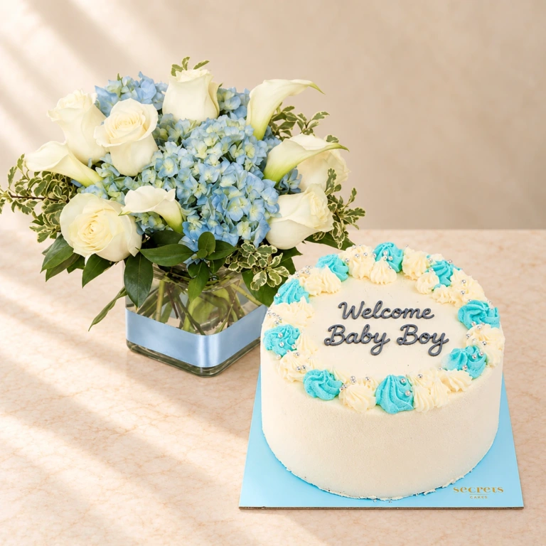 Pale blue hydrangea and white roses in a square vase beside a blue cake reading Welcome Baby Boy